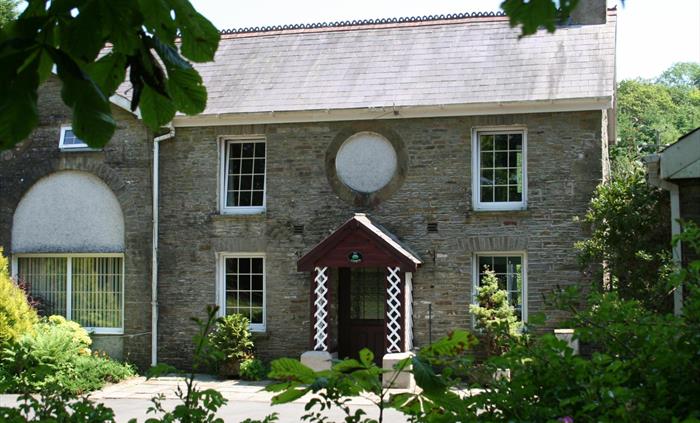 A charming view of Ty Cerbyd’s traditional stone facade with symmetrical sash windows and a decorative circular feature above the door, surrounded by